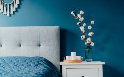 Stylish modern bedroom in dark colors. Cozy interior with navy blue walls, home decor. Bed with gray fabric headboard, blue blanket, bedside table, vase with natural cotton flowers, candles on tray